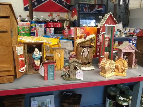 A display of decorative outhouses and figurines on a table, with various products and a Confederate flag in the background.