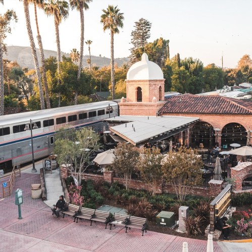 A scenic view of a train station with palm trees, outdoor seating, and a historic building under a clear sky.