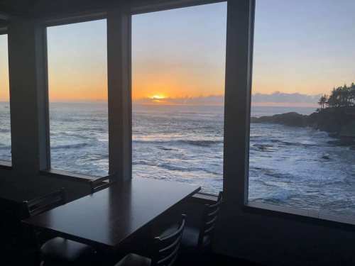Sunset over the ocean viewed through large windows, with a table and chairs in the foreground.