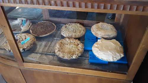 A display case with various homemade pies, including fruit and cream varieties, on a wooden shelf.