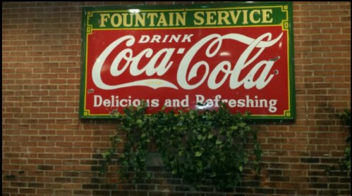 Vintage Coca-Cola sign on a brick wall, featuring "Fountain Service" and "Delicious and Refreshing" with greenery.