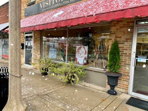 A storefront with a red awning, featuring a holiday display and light snow falling in the foreground.