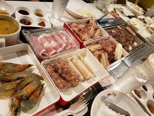 A table set with various raw meats, shrimp, sausages, and dipping sauces for a barbecue meal.