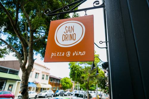 Sign for "San Drino" featuring "pizza & vino," hanging outside in a tree-lined street.