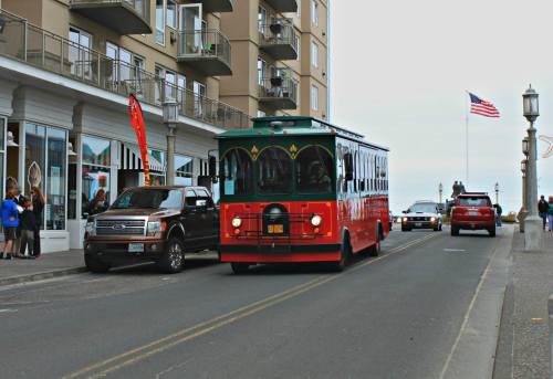 A red trolley bus drives down a street lined with buildings and parked cars, with an American flag in the background.