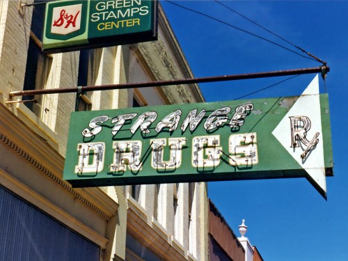 A vintage sign for "Strange Drugs" with an arrow, set against a clear blue sky.