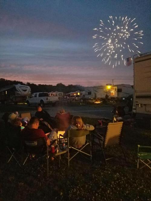 A group of people sitting around a campfire, watching fireworks in the night sky at a campsite.
