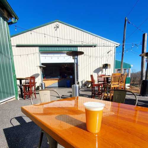 A sunny outdoor seating area with a wooden table and a glass of beer in the foreground, and a building in the background.