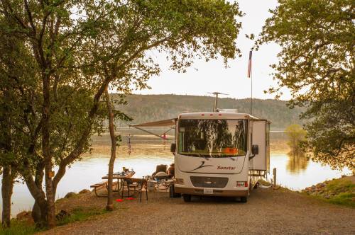 A parked RV by a serene lake, surrounded by trees, with a table set up outside under an awning.