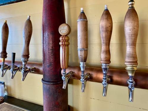 A row of wooden beer taps mounted on a wall, with a red pipe in the background.