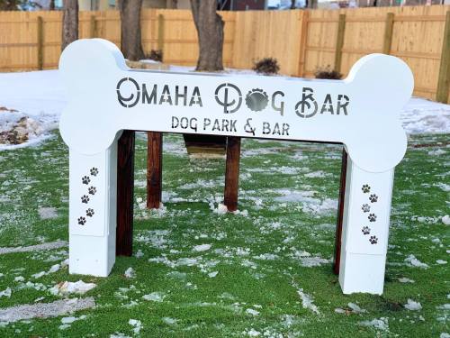 Sign for "Omaha Dog Bar" at a dog park, featuring a large bone shape and paw prints on the sides, with snow on the ground.