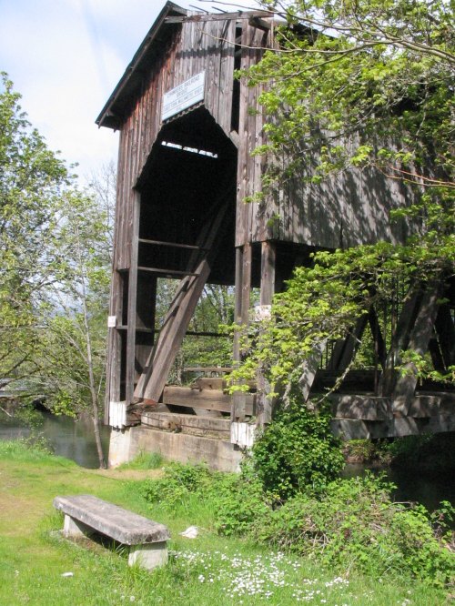 A weathered wooden covered bridge stands over a small stream, surrounded by greenery and a grassy area.