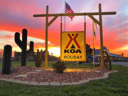 KOA Holiday sign with cacti and rocks, set against a vibrant sunset sky with an American flag in the background.