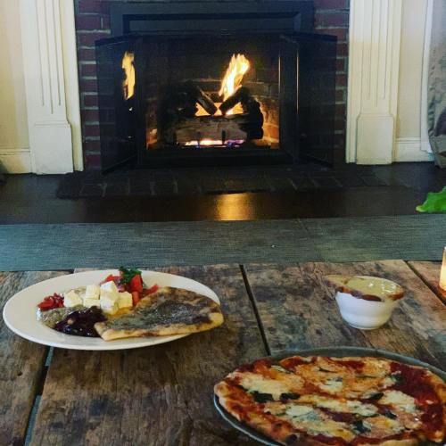 A cozy scene with a fireplace, featuring a plate of food and a pizza on a rustic wooden table.