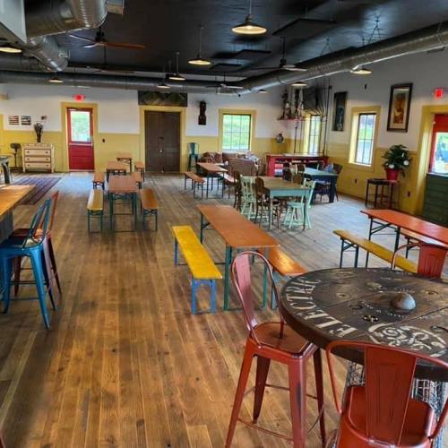 A spacious, brightly lit room with wooden floors, colorful tables, and chairs, featuring large windows and rustic decor.