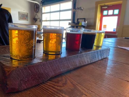 A wooden flight board with five small glasses of different colored beers on a rustic table.