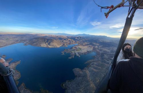 Aerial view from a hot air balloon over a lake and mountainous landscape, with passengers visible in the basket.