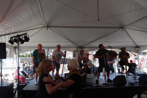 A band performs on stage under a tent while people dance in front of them at a lively outdoor event.