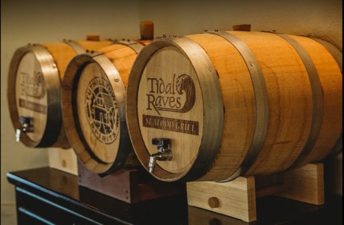 Three wooden barrels with taps, labeled "Tidal Raves" and "Uphill Spirits," displayed on a dark surface.