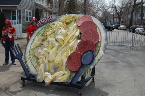 A large, colorful float resembling a plate of haluski with noodles, cabbage, and sausage, displayed on a street.