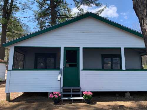 A white cabin with green trim, featuring a porch and flower pots, surrounded by trees under a blue sky.