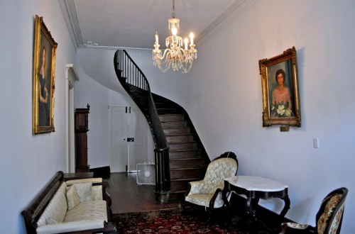 A spacious hallway with a staircase, chandelier, vintage furniture, and framed portraits on the walls.