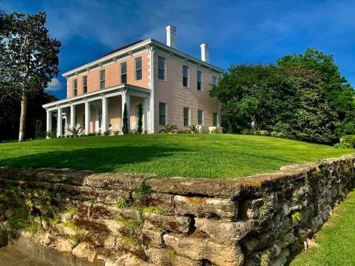 A large, historic house with a pink facade and white columns, surrounded by lush green grass and trees.