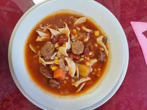 A bowl of hearty soup with noodles, vegetables, and meatballs, served on a white plate against a red tablecloth.
