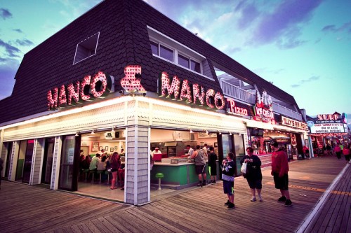 A lively pizza shop with bright signage, bustling with customers on a boardwalk at dusk.