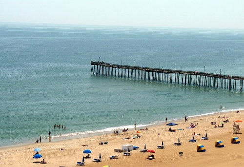 A sandy beach with colorful umbrellas, people swimming, and a long wooden pier extending into calm blue waters.
