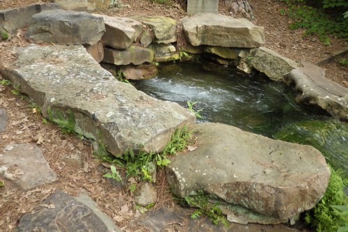 A natural stone pool surrounded by rocks and greenery, with water gently flowing in a serene outdoor setting.