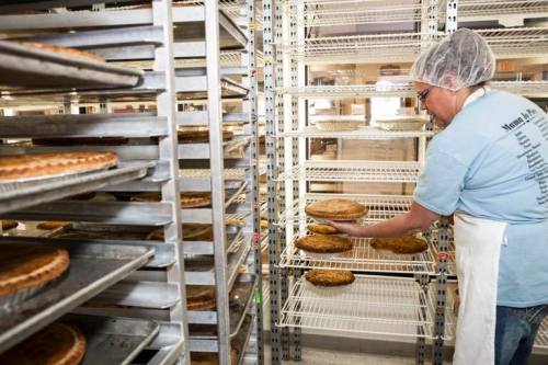 A baker in a hairnet places a pie on a rack in a bakery filled with baked goods.