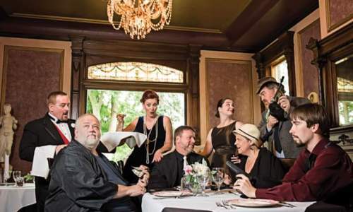 A group of elegantly dressed people gathered around a dining table in a vintage restaurant setting, with a chandelier above.