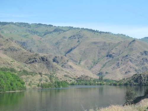 A serene landscape featuring a calm lake surrounded by rolling hills and green vegetation under a clear blue sky.