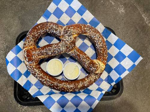 A large, salted pretzel on a blue and white checkered paper, accompanied by two small cups of dipping sauce.