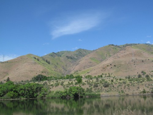 A serene landscape featuring rolling hills, green vegetation, and a calm body of water under a clear blue sky.
