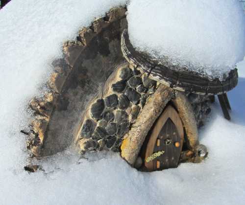 A whimsical miniature house made of wood and stones, partially covered in snow.