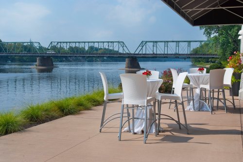 Outdoor dining area by a river, featuring white tables and chairs, with a green bridge in the background.