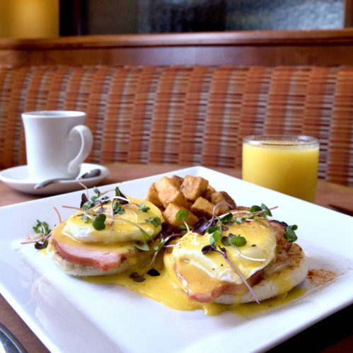 A plate of eggs Benedict with ham, hollandaise sauce, and microgreens, served with potatoes and drinks.