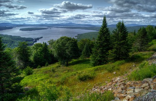 A scenic view of a river surrounded by lush greenery and mountains under a cloudy sky.