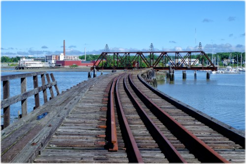 A weathered wooden bridge with railway tracks extending over calm water, under a clear blue sky.