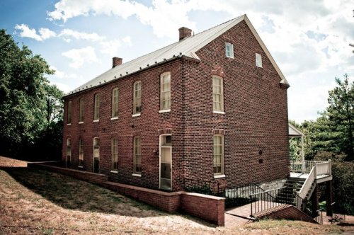 A historic brick building with a sloped roof, surrounded by greenery and a staircase leading to a lower level.