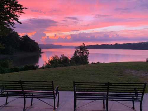 A serene lakeside view at sunset, with vibrant pink and purple skies reflecting on the water, and two empty benches.