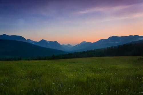 A serene landscape at dusk, featuring rolling mountains and a grassy field under a colorful sky.
