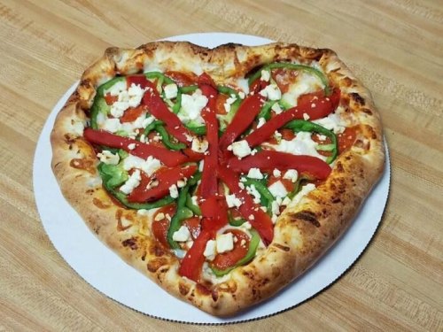 A heart-shaped pizza topped with green peppers, tomatoes, and feta cheese on a wooden surface.