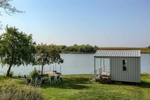 A serene lakeside scene featuring a small cabin, a dock, and seating area surrounded by greenery under a clear sky.