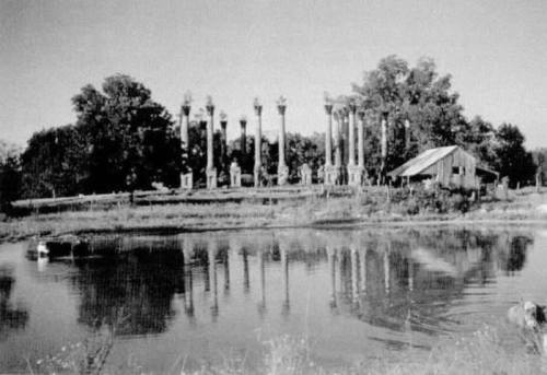 Black and white image of a pond with a barn and tall columns in the background, surrounded by trees.