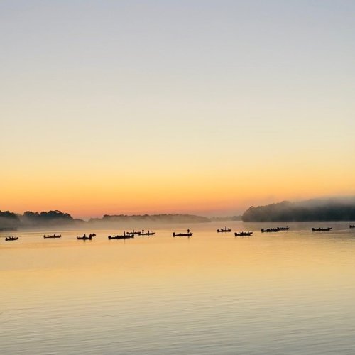 A serene lake at dawn, with mist rising and several boats gently floating on the calm water under a colorful sky.