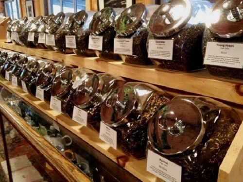 A row of glass jars filled with various loose teas, labeled and displayed on a wooden shelf.