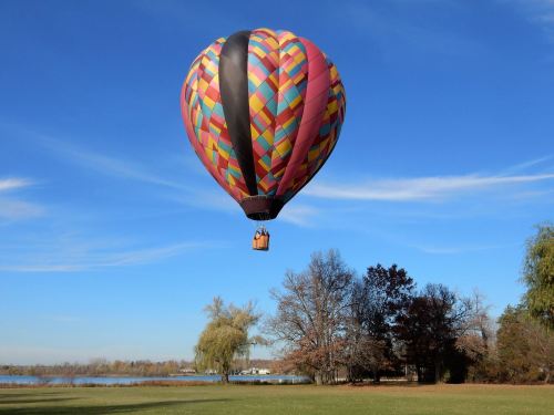 A colorful hot air balloon floats above a grassy area near a calm lake under a clear blue sky.